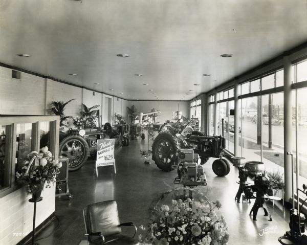 Agricultural equipment on display among potted plants and flower arrangements in baskets. The display is in the showroom of International Harvester's Indianapolis Branch House. A row of large windows are on the right, which borders a sidewalk and street with automobiles parked near the curb.
