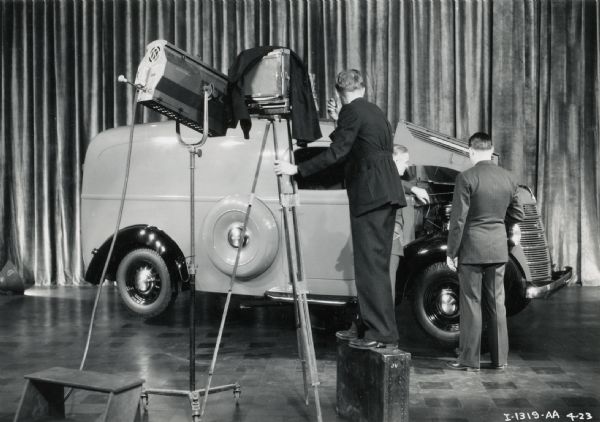 A photographer uses a view camera to photograph an International truck placed in front of a curtain. Two other men are standing near the open hood of the vehicle.