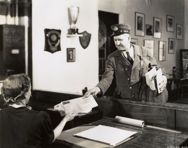 A postman handing an envelope to a woman sitting behind a desk in an office. The woman is wearing a headset, perhaps for a telephone.