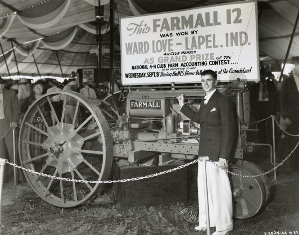 Ward Love, winner of a Farmall-12, stands next to his prize in a tent at the Indianapolis State Fair.  The sign on the tractor reads: "This Farmall 12 was won by Ward Love - Lapel, Ind. 4-H Club Member as Grand Prize of the National 4-H Club farm Accounting Contest.... To Be Presented to Him Wednesday, Sept.8th During the WLS Dinner Be...at the Grandstand."