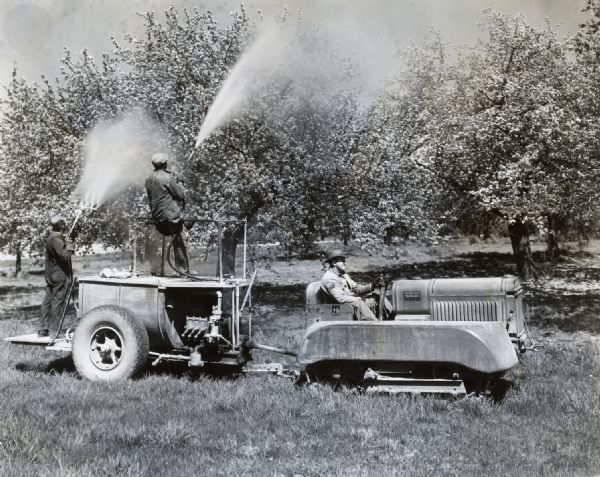 Three men use an International T-20 TracTracTor (crawler tractor) with orchard fenders and a 300-gallon bean spray tank. The men are applying pesticide(?) to fruit trees in an orchard.