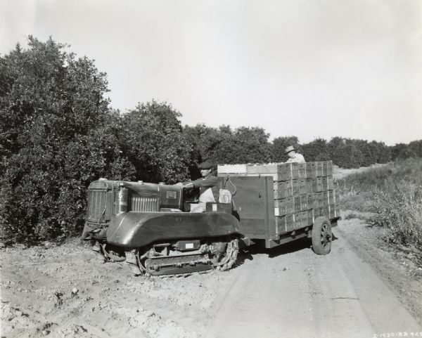 Two men use an International T-20 TracTracTor (crawler tractor) to haul orange boxes on a trailer from a grove in an orchard to be loaded onto trucks. The tractor is equipped with orchard fenders. The original caption reads: "Owner: --Western Fruit Growers; Address: --Redlands, California; Location of Job:--Between Redlands and Yucaiapa; Model:--T-20; Bought:--October, 1935; Description of work:--Picking up and hauling orange boxes from grove to where they can be loaded onto trucks. Small trailers hold 80 boxes averaging 56 pounds, or 4,480 pound loads.; Loads or trips per day:--Approximately 50; Other work performed:--Also used for ditching cultivating; Fuel:--Gasoline; Hourly consumption:--1.6 gal."