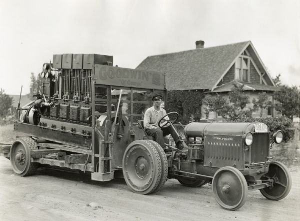 A man sits behind the wheel of a McCormick-Deering 10-20(?) tractor attached to a trailer with a sign reading: "Goodwin's: Of Course." There is a building in the background.