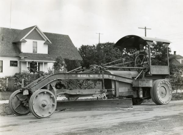 A man stands behind the wheel of a road grader marked: "Russell Motor Patrol No.3." The grader is on a residential street with a house in the background.