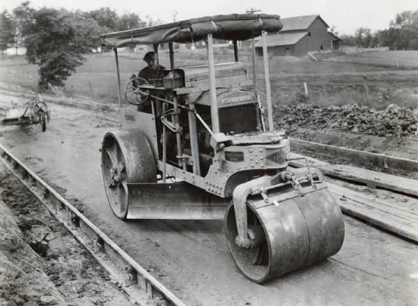 A man operates a McCormick-Deering 10-20 tractor with an Acme Road Roller to work on United States Road 63. There is a barn in the background.