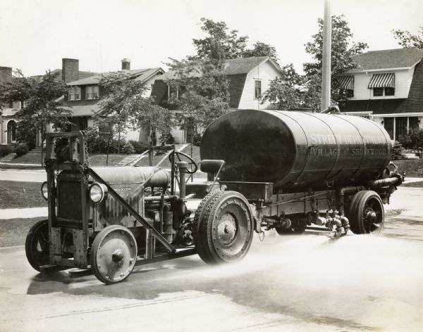 McCormick-Deering 10-20 tractor pulling a street cleaning tank attachment marked "Street Department, Village of Shorewood." Water is spraying from the side of the tank onto the road. Houses are in the background.