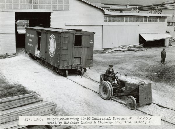 A man uses a McCormick-Deering 10-20 industrial tractor to pull a boxcar out of a building. The boxcar is running on railroad tracks. The tractor was owned by Hutchins Lumber and Storage Company.