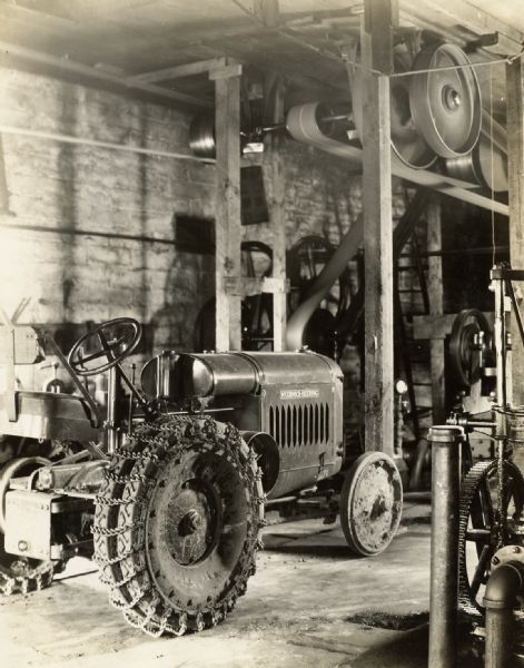 A McCormick-Deering 10-20 industrial tractor outfitted with tire chains sitting parked in a garage or factory. There is belt-driven machinery near the ceiling.