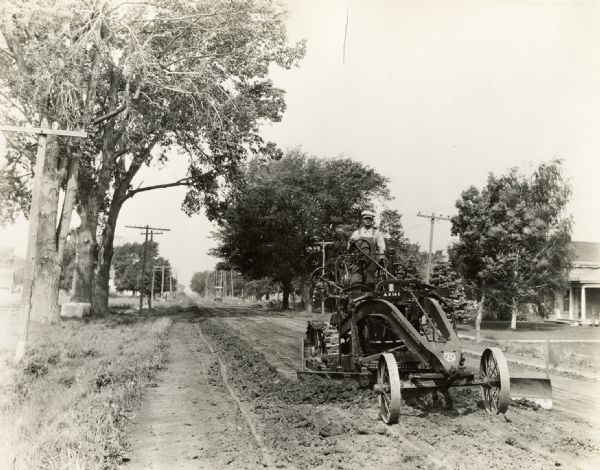 A State Highway Commission worker operates an Austin Western motor grader on Road #13 from Webster to South English in Keokuk County. There is a house in the background.
