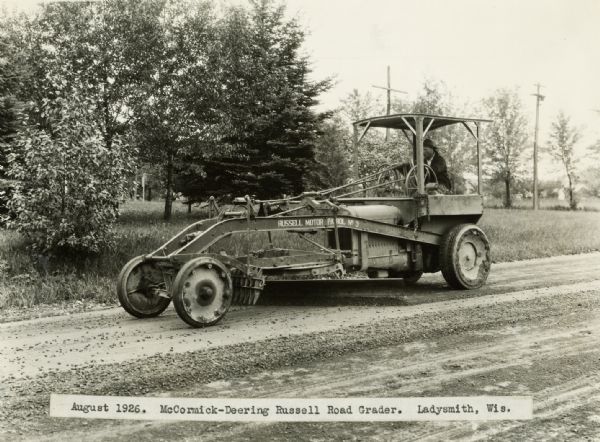 A man operates a Russell road grader on a rural road.