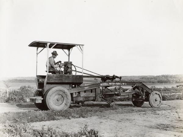 A man operates a grader marked "Russell Motor Patrol No.3" on a rural road.