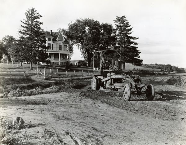 Barto Speer operates a grader marked "Russell Motor Patrol No.3" on a rural road in Dallas County, Iowa. There is a large house and lawn in the background.
