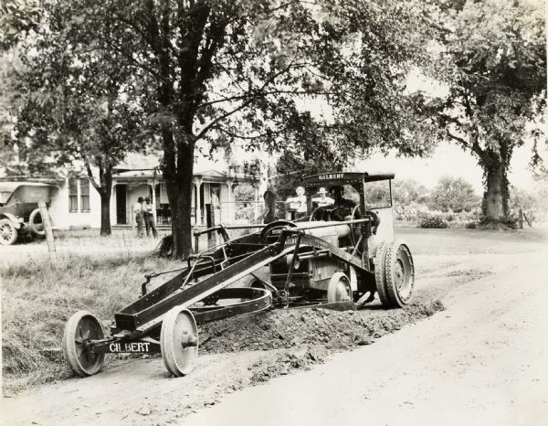 W.H. Stuber operates a Gilbert road grader on a road in "Northern Park." Men are standing in the background near a house.