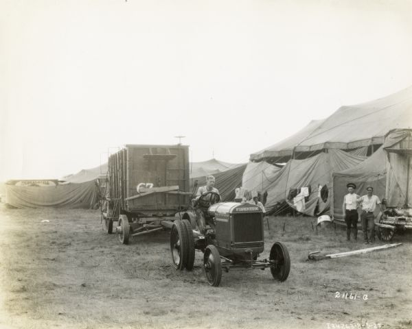 A man pulls a circus wagon with a McCormick-Deering 10-20 tractor while two men watch near a tent. The circus is likely the Sells-Floto Circus.
