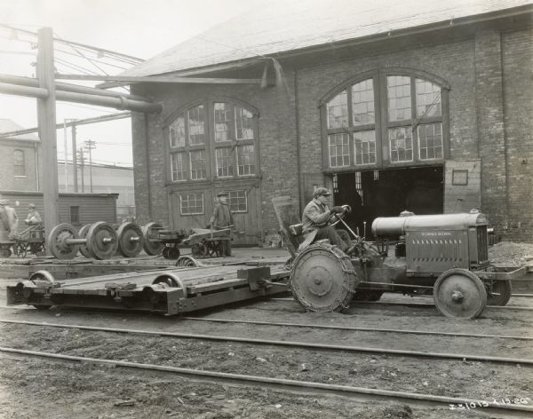 A worker uses a McCormick-Deering 10-20 tractor to pull a platform along tracks in a railroad yard outside a factory or other type of industrial building.