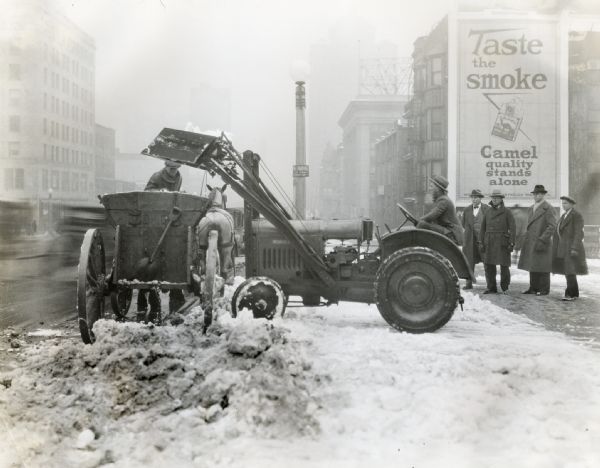 A man using a McCormick-Deering 10-20 tractor to load snow from a city street into a horse-drawn wagon. A group of men are watching from the sidewalk, and a Camel cigarette billboard is in the background.