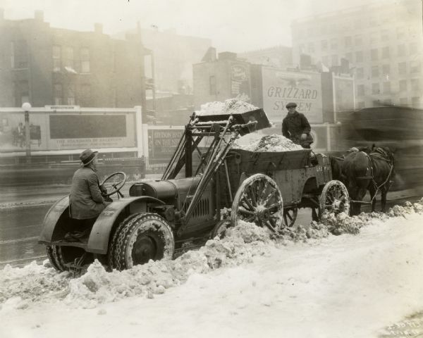 Man using a McCormick-Deering 10-20 industrial tractor to load snow into a horse-drawn wagon on a city street. A billboard advertising the "Grizzard System" is in the background.