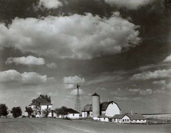 Farmhouse with several barns, a silo, and a windmill surrounded by fields and a bright, cloud strewn sky.