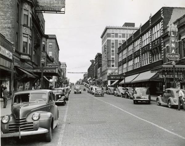 Automobiles driving down Main Street, which is lined on either side with storefronts and buildings. On the left side of the street is Hotel LaFayette. On the right side is the Luna Theatre, Western Union, Hotel Kankakee, and Lassers Furniture. A banner hanging over the street reads: "Sears Anniversary Sale."