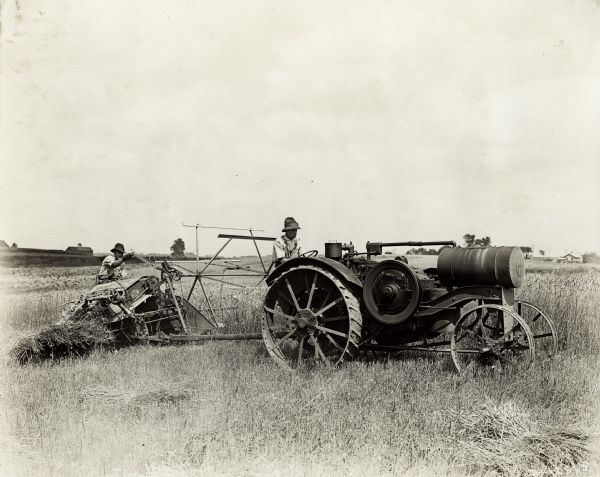 A man pulling an Osborne grain binder with a Titan 10-20(?) tractor.  In the background are farm buildings. Bundled piles of grain are in the lower right corner.