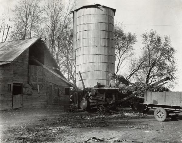 A man works with a husker-shredder near a silo and barn.  A wagon is parked on the right.  Original caption states:  "Husker and Shredder used for custom work by A-M. Motor and Implement Company."