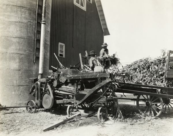 Two men are working with a husker-shredder near a silo and barn. One of the men is shoveling corn stalks from a wagon.