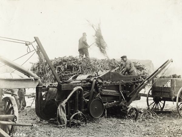 A man is standing on a wagon loaded with corn stalks while another man is operating a husker-shredder.