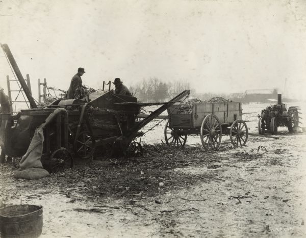 Men loading corn stalks from a wagon into a husker-shredder in a snow-covered field. A second wagon and a tractor are on the right, and there is a barn in the background.