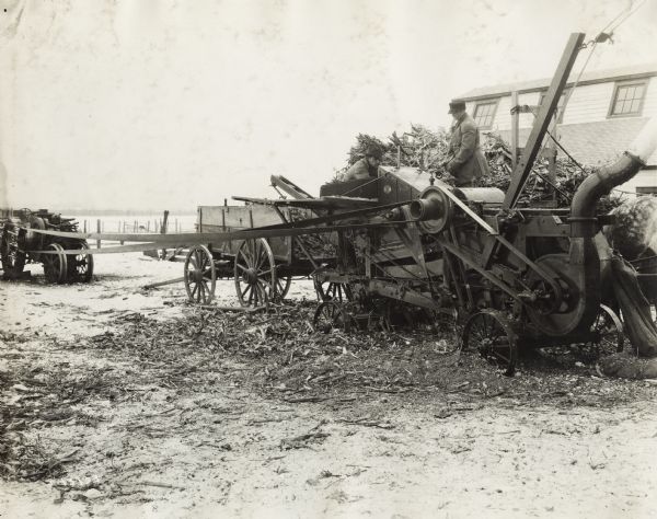 Two men loading corn stalks into a husker-shredder from a horse-drawn wagon in a snow-covered field.  A building with windows near a sloped roof is behind them.  A belt connects the husker-shredder to the power-takeoff of a tractor.