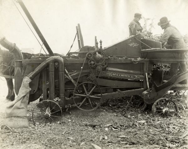 Two men are feeding corn stalks from a horse-drawn wagon into a McCormick husker-shredder.
