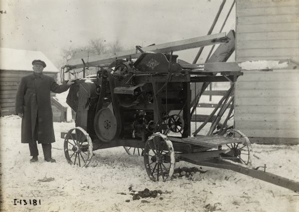 A man in a hat and knee-length coat stands next to a husker-shredder.  Several farm buildings and trees are in the background, and snow is on the ground.