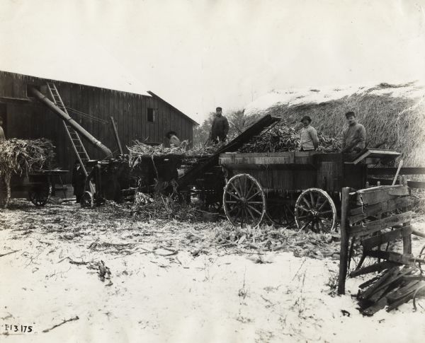 A crew of men standing with a wagon and a husker-shredder near a barn. A sixth man in a dark coat and hat is standing under the ladder leaning against the barn. Corn and husks fill the wagons and cover the ground.  A large pile of hay is on the right. Both the hay and the barn are covered with snow and icicles are hanging from the barn.