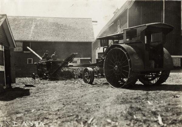Two men working with a husker and shredder in a barnyard. The machine is connected with a belt to the power-takeoff of a Mogul tractor. Original caption states: "Husking corn at the rate of 85 bushels per hour and making nutritious stores(?) from the stalks with a McCormick Improved Husker and Shredder operated by an I.H.C. Oil Engine."