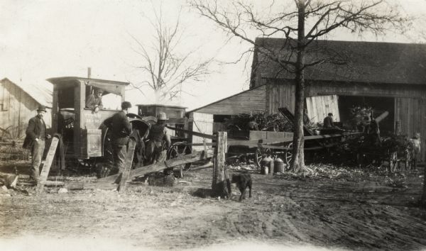 Men working with wagons and husker-shredder near a barn. The husker-shredder is powered by a Titan 18-35 tractor. A dog is walking by a fence, and milk cans are next to a tree.