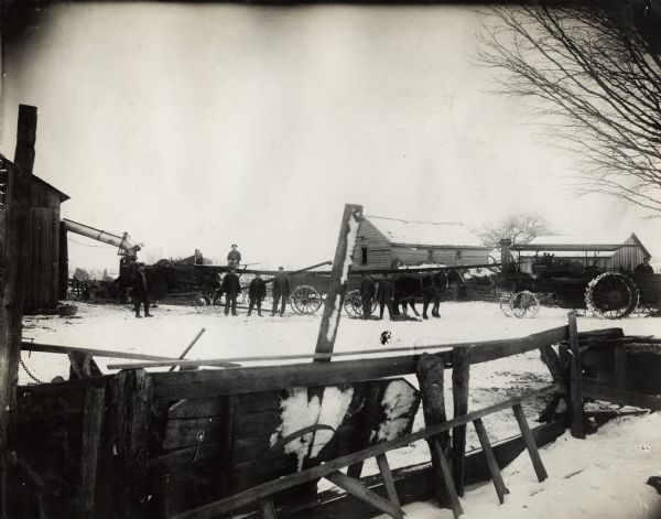 Six men are standing by a husker-shredder, wagon, and two horses in a farm yard. A tractor, possibly a Mogul 8-16 is on the right. Barns and farm buildings surround the yard in which the men are standing. A fence is in the foreground and snow covers the ground and buildings.