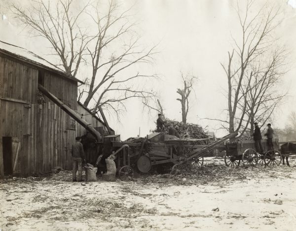 Two men are working with a husker-shredder, one moving corn from a wagon, the other feeding them into the machine. Two other men are at a wagon on the right, which is filled with corn, and has a horse hitched to the front. On the left near some sacks a man is standing and watching the operation. Snow is on the ground and on the roof of the barn.