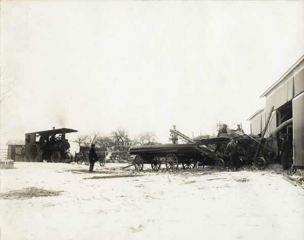 Several men are working around a husker-shredder and a wagon loaded with corn. A barn is one the right. The husker-shredder is attached to a tractor. A fence, wagon, and trees are in the background. Snow covers the ground.