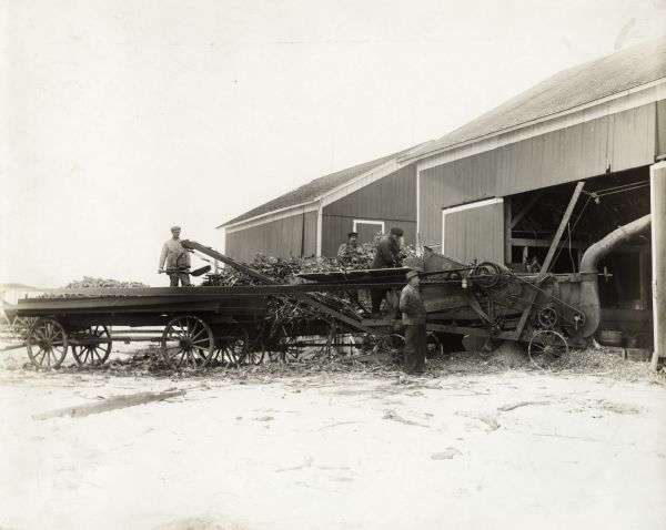 Men feed corn from two wagons into a husker shredder near a barn in a snow-covered farm yard. Original caption states:  "Owned and operated by Wm. Stiver & Sons."