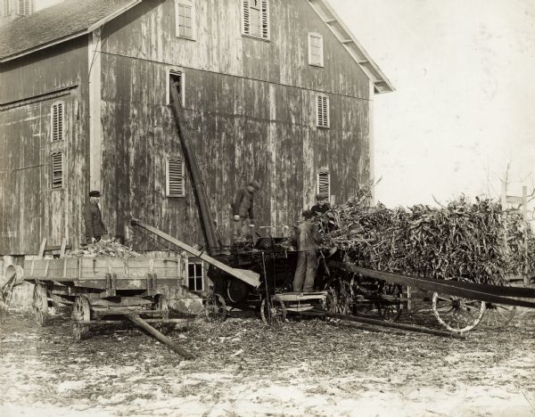 Men feed corn from wagons into a husker-shredder. The men are working near a barn in a snow-covered farm yard.  Original caption states:  "Owned and operated by C.L. Troup, Milo Gyeer. Jess Eisenhour, Victor Fuller of New Paris, Ind.  Sold by Weybright Bros., New Paris, Ind.  McCormick Little Giant Husker and Shredder operated by Mogul 8-16 (South Bend Territory)."