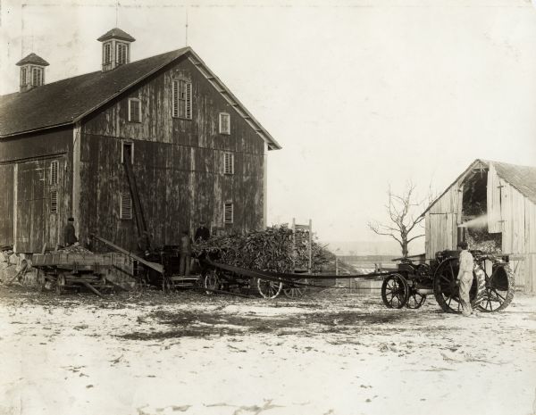 Three men loading corn into a husker-shredder near a barn. The husker-shredder is connected to a Mogul 8-16 tractor. Original caption states:  "The 8-16 shown in this picture is owned by Weighbright Bros., New Paris, Ind. Shredder owned and operated by C.L. Troup, Milo Gyeer, Jess Eisenhour, and Victor Fuller. (South Bend Territory)."