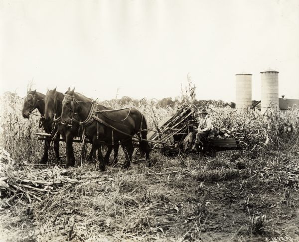 Farmer operating a horse-drawn corn binder in a field. Two silos and a barn are in the background.
