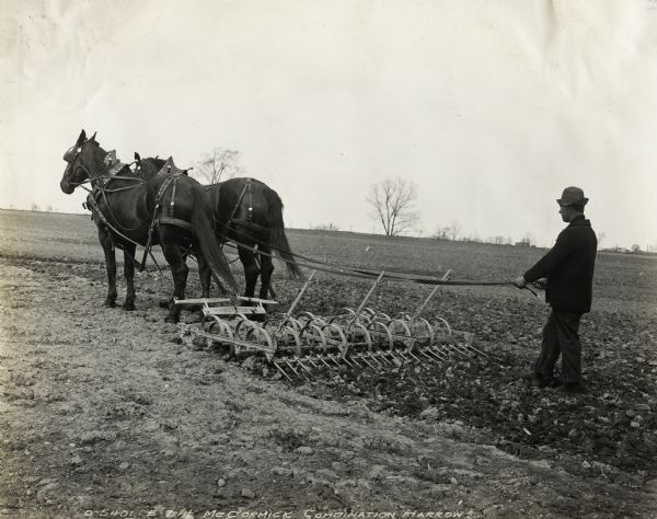 A farmer walks behind a horse-drawn combination harrow. A fence, trees, and buildings are on the horizon.