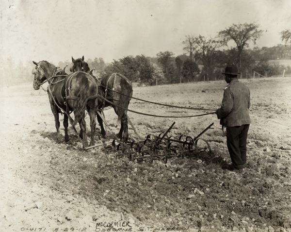 Farmer wearing a hat is walks=ing behind a horse-drawn Osborne spring tooth harrow in a field. Trees and a fence are in the background.