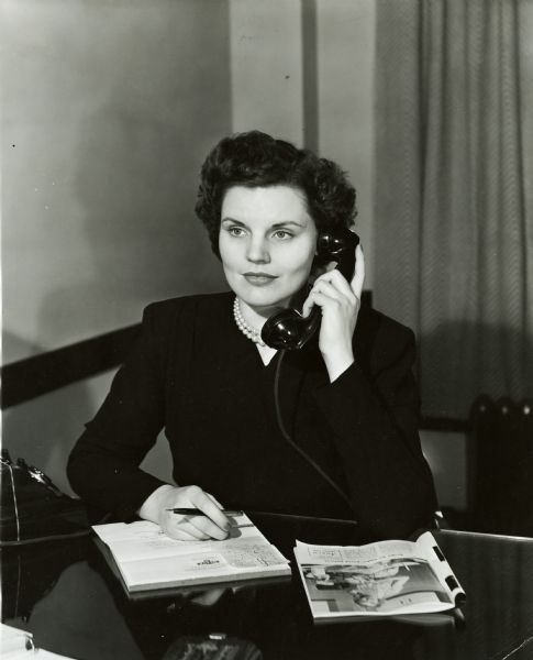 Miss Olive White of the Home Economics Department at International Harvester's Evansville Works. She is sitting at a desk, with a phone to her left ear. A magazine sits open next to a pad of paper.