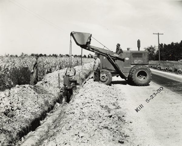 A crew of men laying sewer pipe with a Hough payloader. Original caption states: "Hough Payloader laying sewer pipe sections weighing 200 lbs each. Equipment owned by Cook County Highway Dept."
