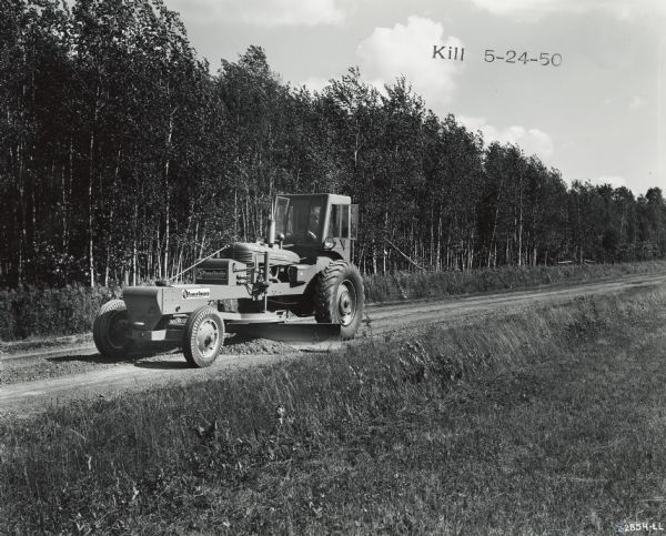 Man spreads gravel along a road with a Meili-Blumberg (MB) Model 10 power grader attached to an International M tractor. One side of the road is lined with trees, the other side is grass. Original caption states: "With this power grader, which has a 12-foot blade and is powered by an International M tractor engine, the town's 48 miles of roadway get a thorough working over every two weeks. Lewis Posekany is board chairman of the Board of Supervisors."