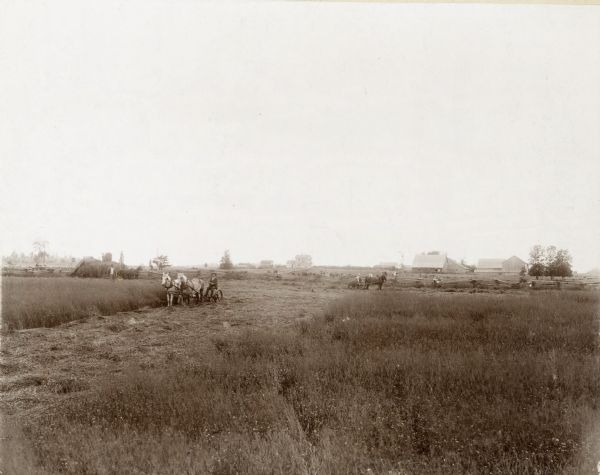 A man is using what appears to be a mower drawn by two horses to work in a field.  Farm buildings and other workers are in the background, as well as children sitting on a fence.