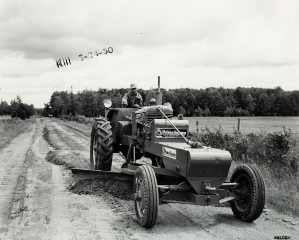 Man grading a dirt road with a Meili-Blumberg Model 10 power grader attached to an International M tractor. Original caption states:  "To maintain its 67 miles of dirt and gravel roads, the Board of Highway supervisors, of which Edward Johnson of Prentice is chairman, recently purchased the MD Model equipped with 12-foot blade." "The grader is powered by an International M tractor engine."