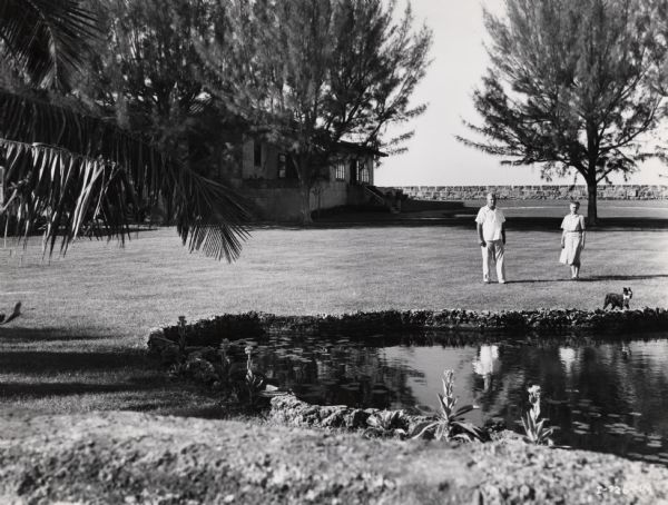 Mr. Heath, manager on an International Harvester sisal plantation in Cuba, walks with his wife in the yard of his official company residence. The couple's dog stands near the pond.