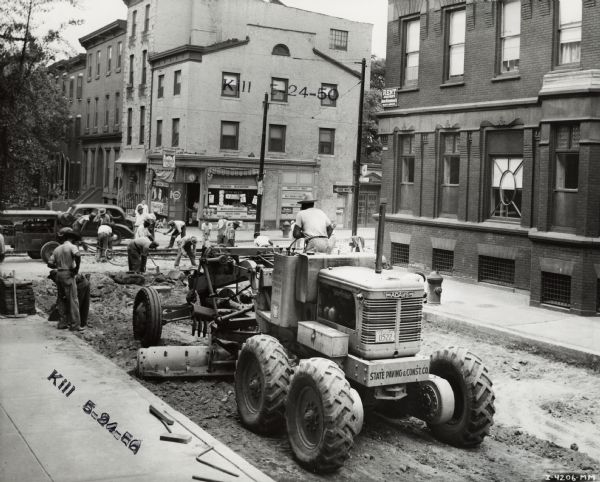 Men using an Adams #5 1/2 Road Grader to work on an urban street. The grader was owned by the State Paving and Construction Co. Original caption states: "Grading street for paving. 22100 block on Spruce St.  Average also 2 blocks a day, rough and fine grading."
